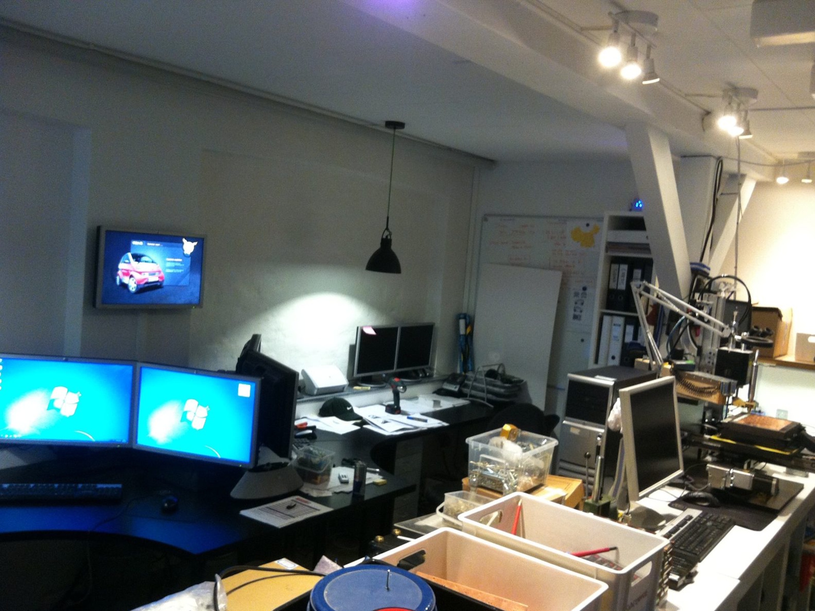 Desks, monitors, parts boxes and a small CNC machine in the office next to the garage, with the electric car on the wall screen