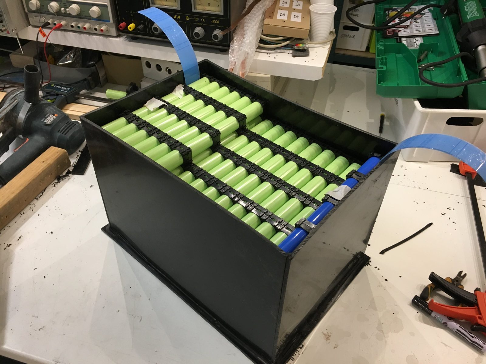 A lithium battery module being assembled on a workbench, rows of green cylindrical cells in black holders inside a black box, with bench power supplies and tools around it