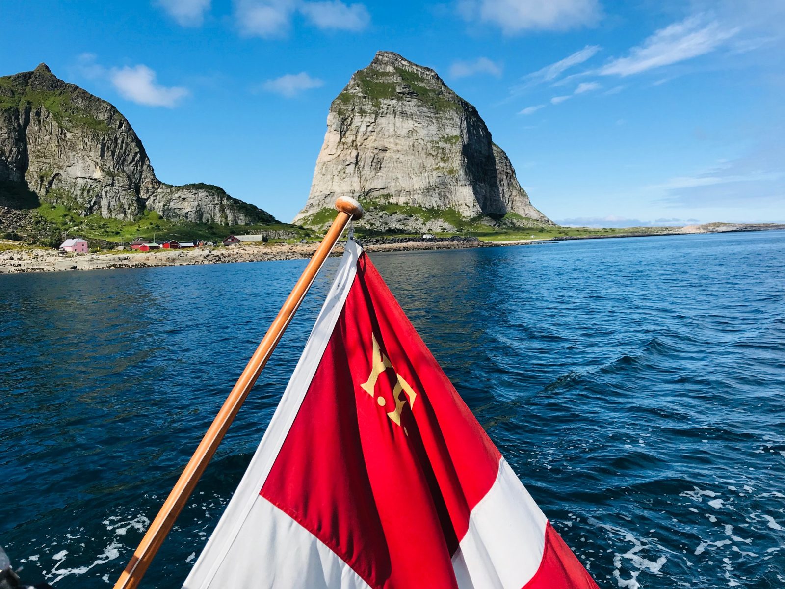 A Danish yacht ensign at the stern, with a steep rock island and red houses on the Norwegian coast behind it