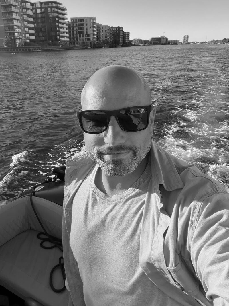 Anders in a small boat on Copenhagen harbour, with apartment buildings and a wake behind him