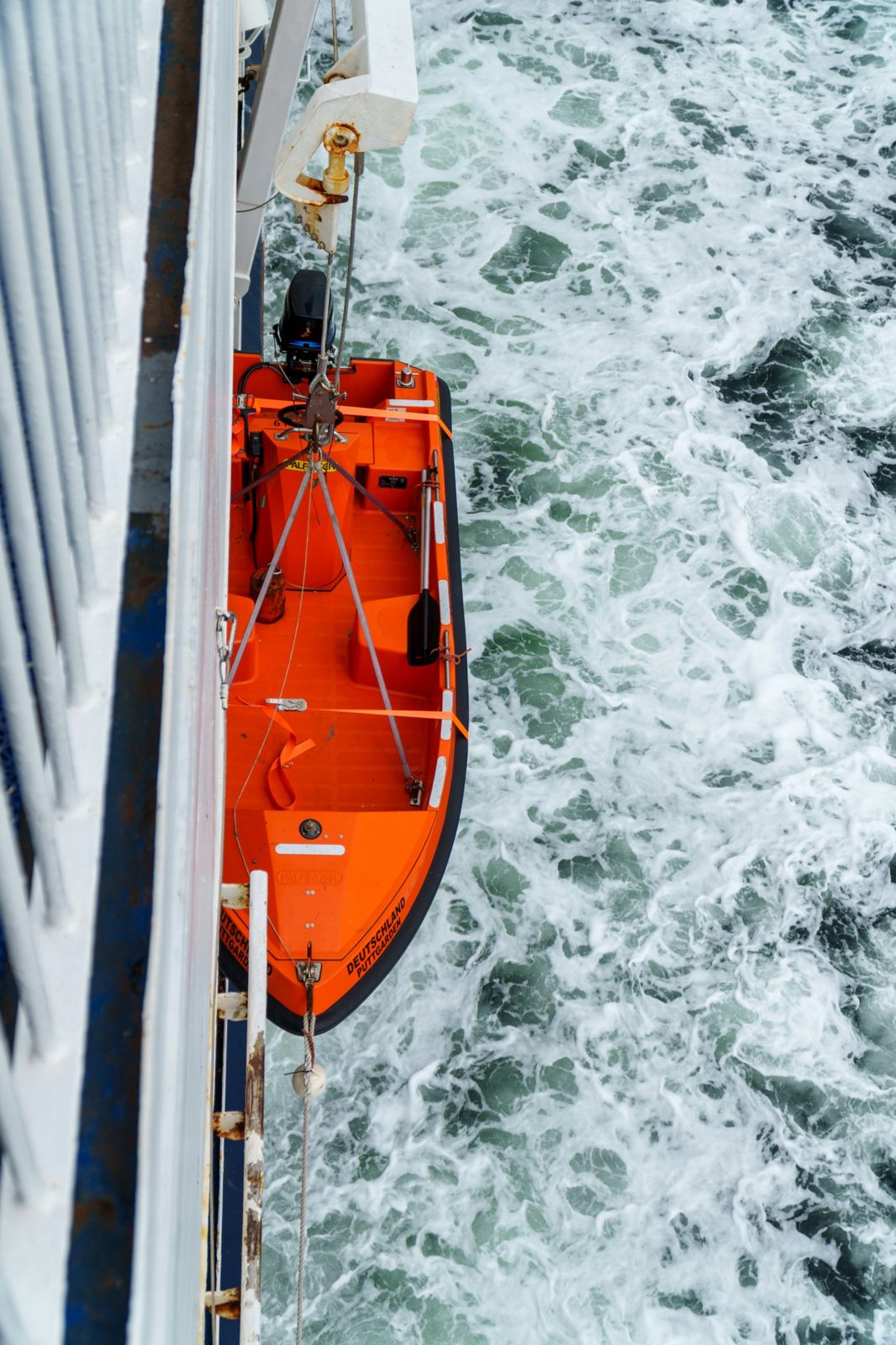 An orange rescue boat hanging in its davit on the side of a ferry, seen from above, with the ship's wake churning below