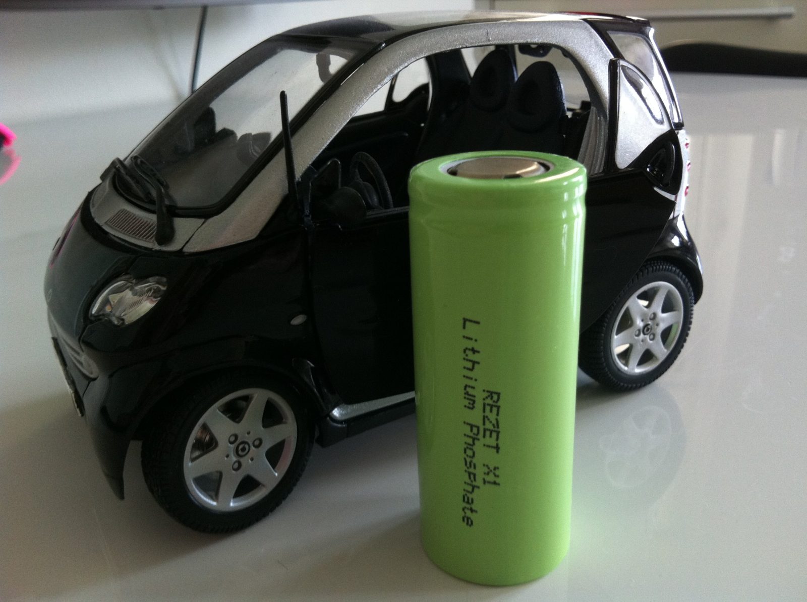 A green lithium iron phosphate cell marked 'Rezet X1' standing next to a model of a small two-seater city car, almost as tall as it