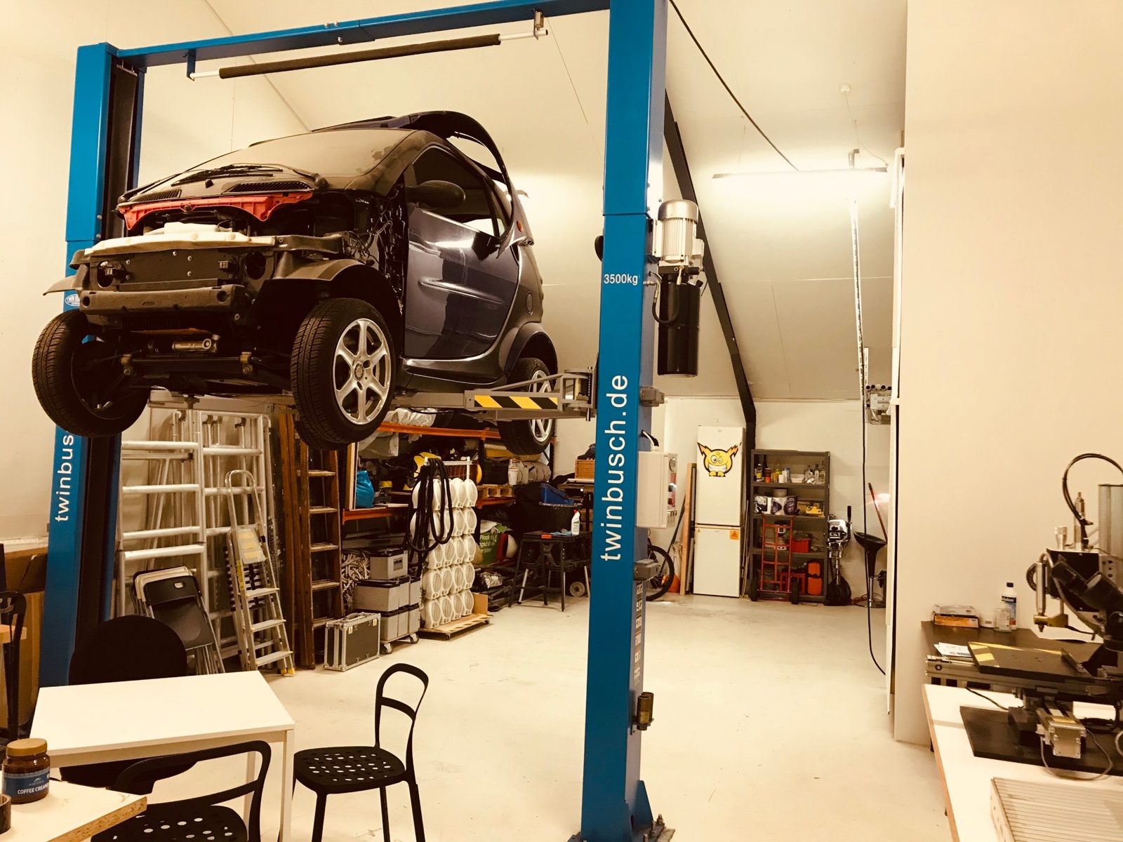 A small car on a two-post lift in a white workshop with shelves and tools